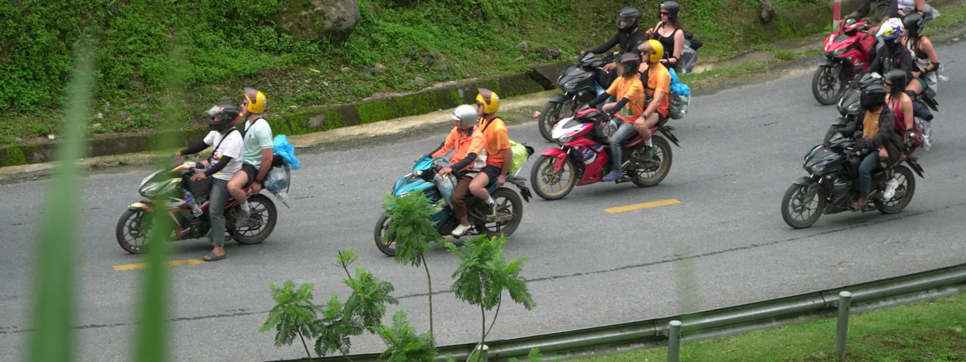 A group of riders on motorbikes travelling along a two-lane mountain road in Hà Giang with a metal crash barrier on the outside of the bend and a steep vegetated bank above the inside