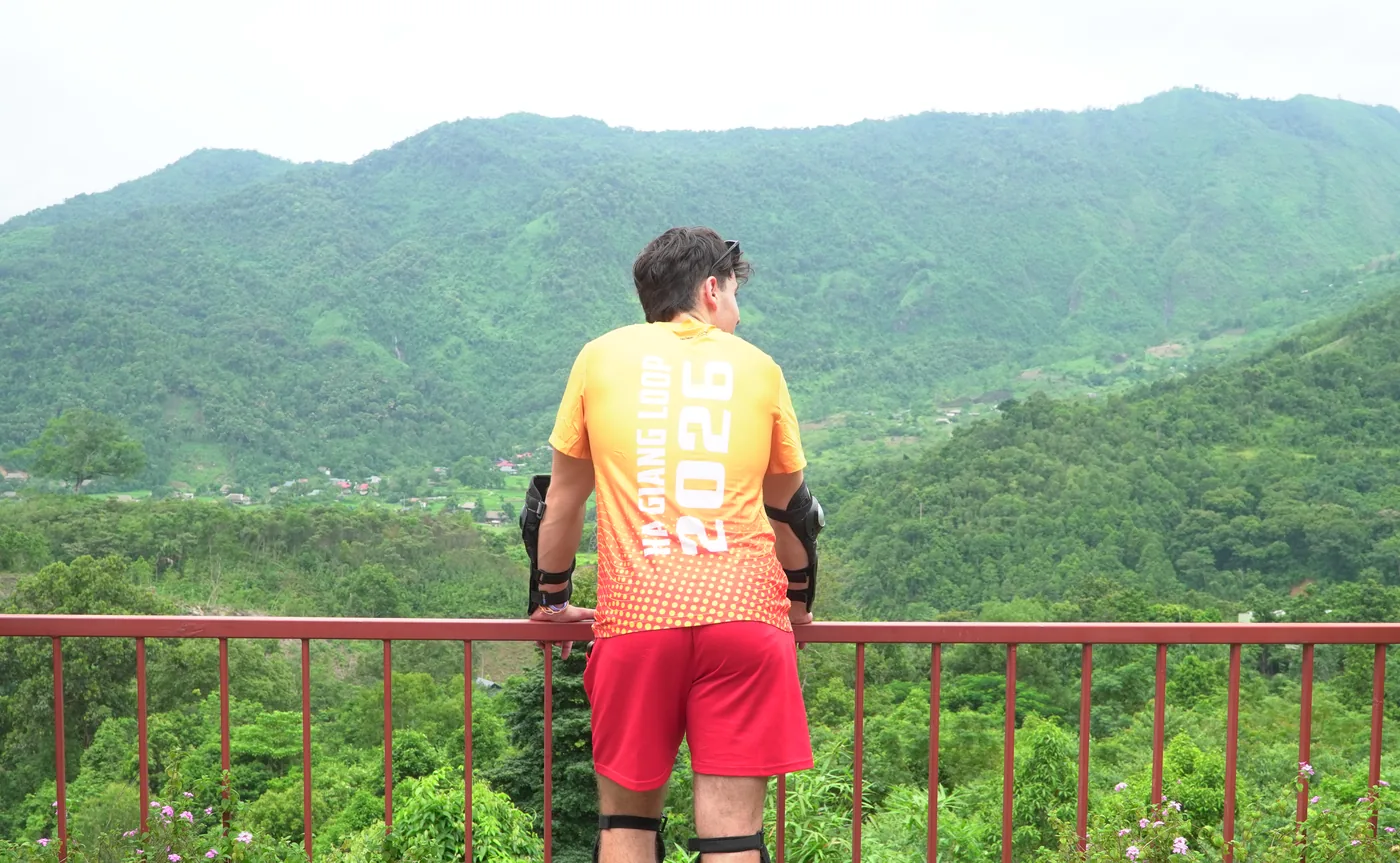 A person seen from behind and slightly to the side at a metal railing, wearing an orange T-shirt printed HA GIANG LOOP 2026 down the back, red shorts and strapped knee pads, looking out over a deep green valley with a scattering of houses and small fields on the far slope under a pale sky