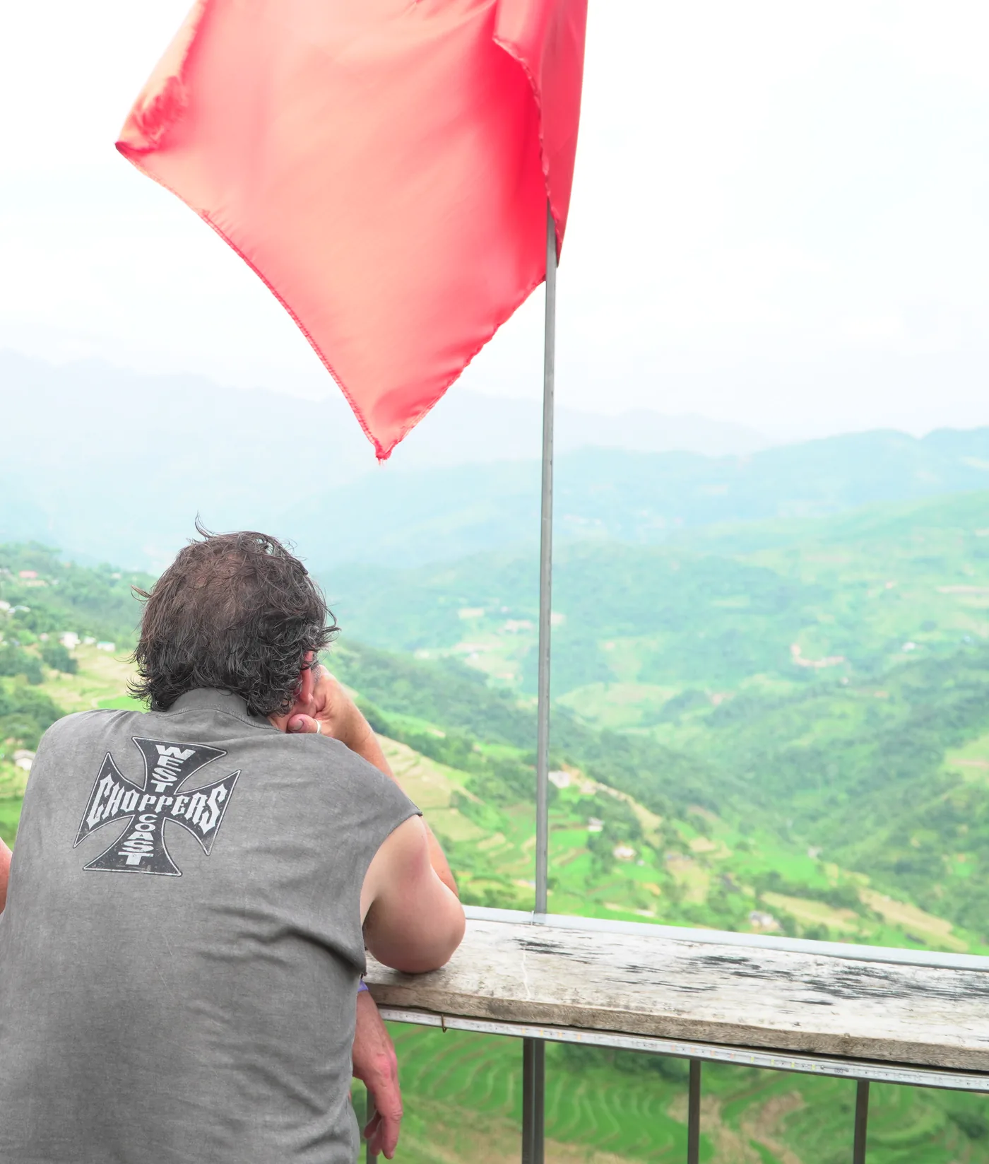 A person seen from behind in a grey sleeveless top with a large cross-shaped graphic on the back, leaning on a weathered timber rail and looking out over a wide valley of green terraced hillsides, scattered houses and hazy ridges. A red flag on a metal pole hangs across the upper part of the frame