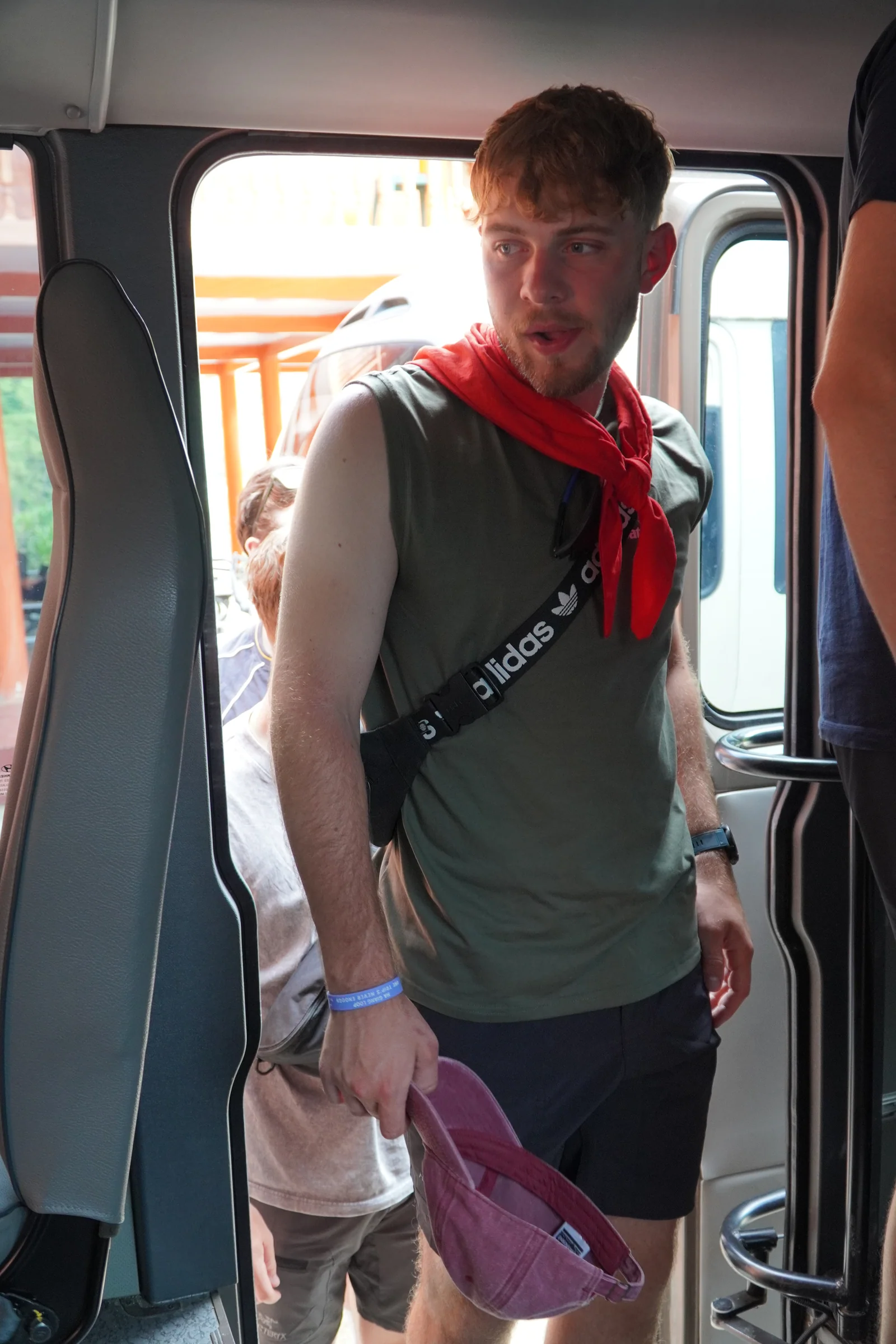 A traveller stepping down out of a sleeper bus doorway with a bag over one shoulder and a red neckerchief, other passengers behind him.