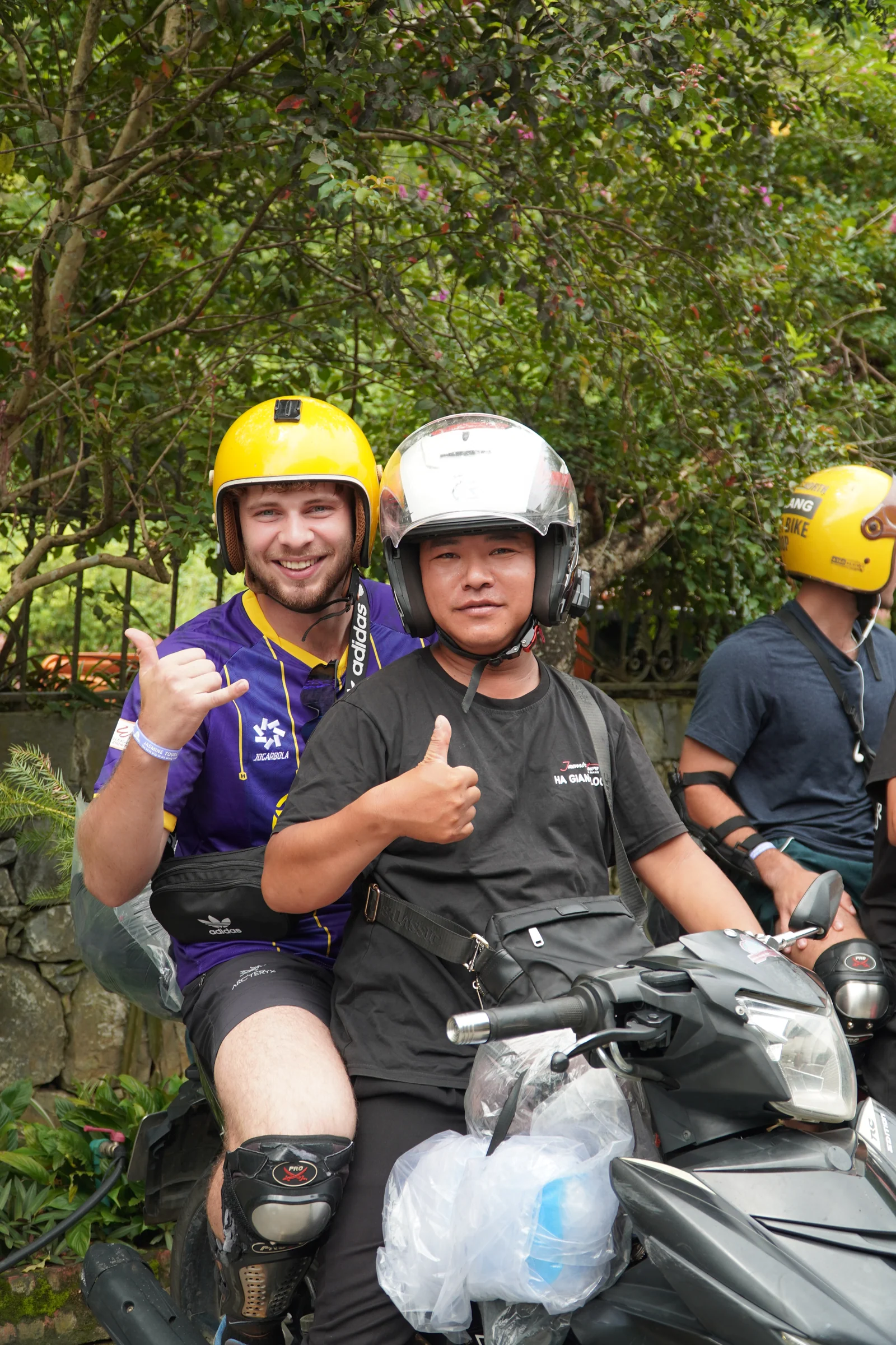 A local driver in a helmet seated on a motorbike giving a thumbs up, a guest in a helmet and body armour sitting behind him doing the same, a second driver waiting behind.