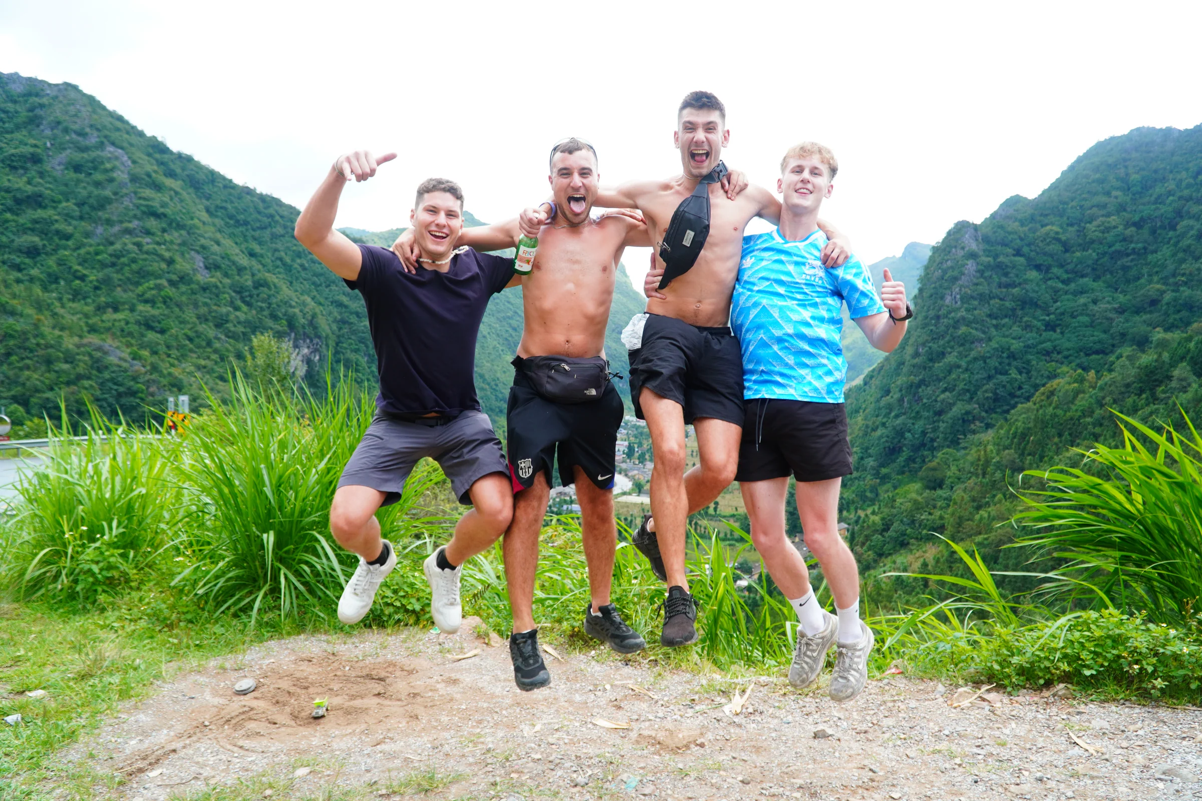 Four travellers jumping in the air together at a roadside viewpoint, arms raised, karst peaks and low cloud behind them.