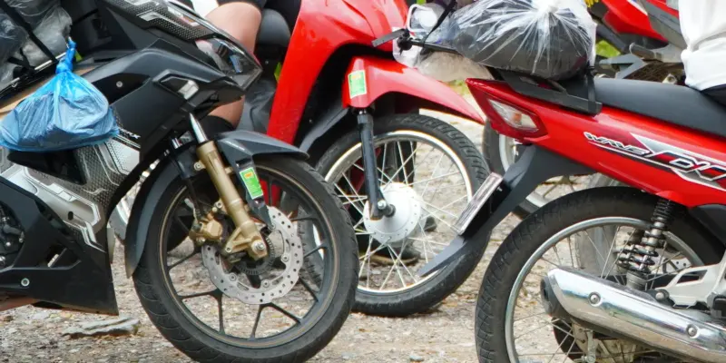 Five motorbikes packed close together on gravel, luggage strapped across the seats and wrapped in plastic bags, a conical hat resting against one load. Two riders are visible from the shoulders down; the frame is cropped so that no face appears.