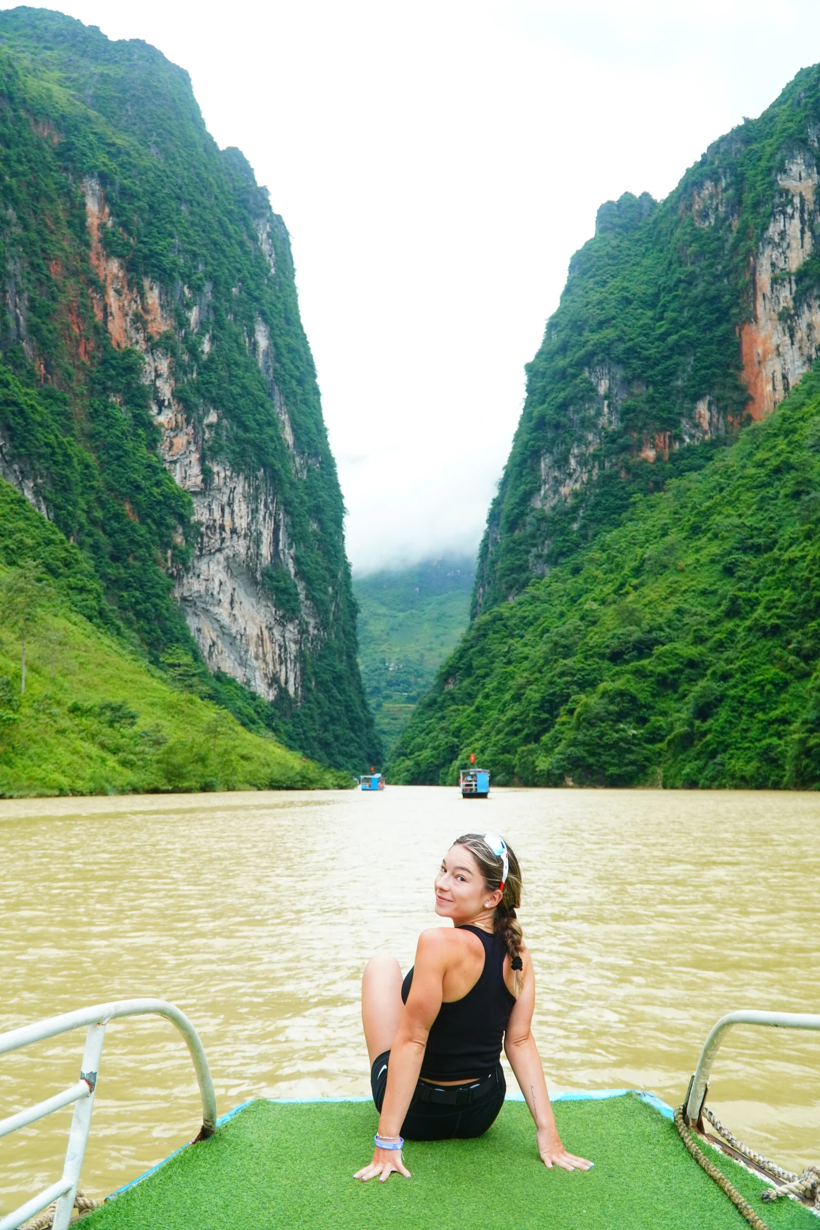 A traveller sitting on the green bow deck of a small boat on the Nho Que river, jade-green water ahead and the limestone walls of the Tu San canyon rising steeply on both sides.