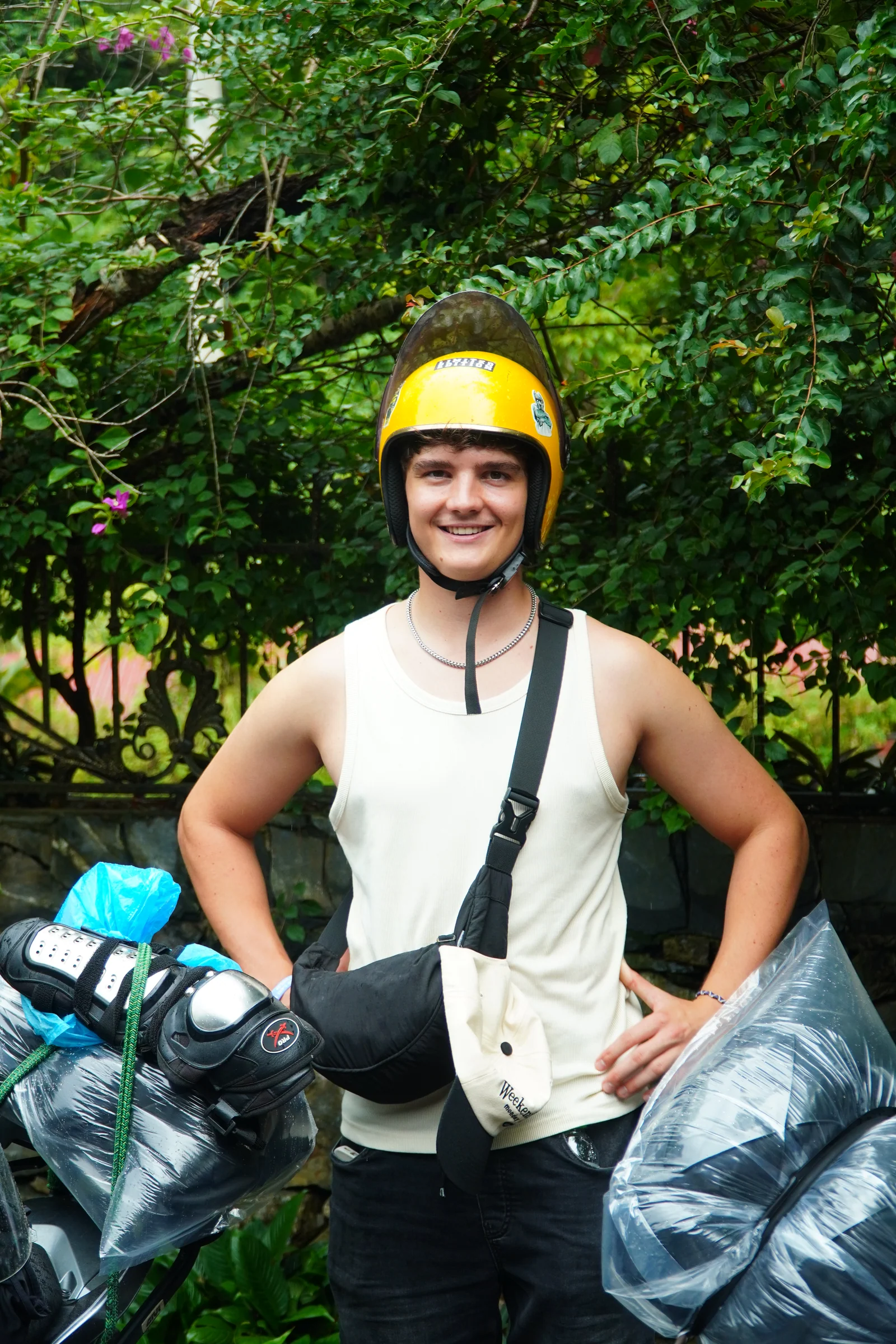 A traveller in a yellow helmet standing beside a bike, holding a folded rain poncho and gloves, greenery behind.