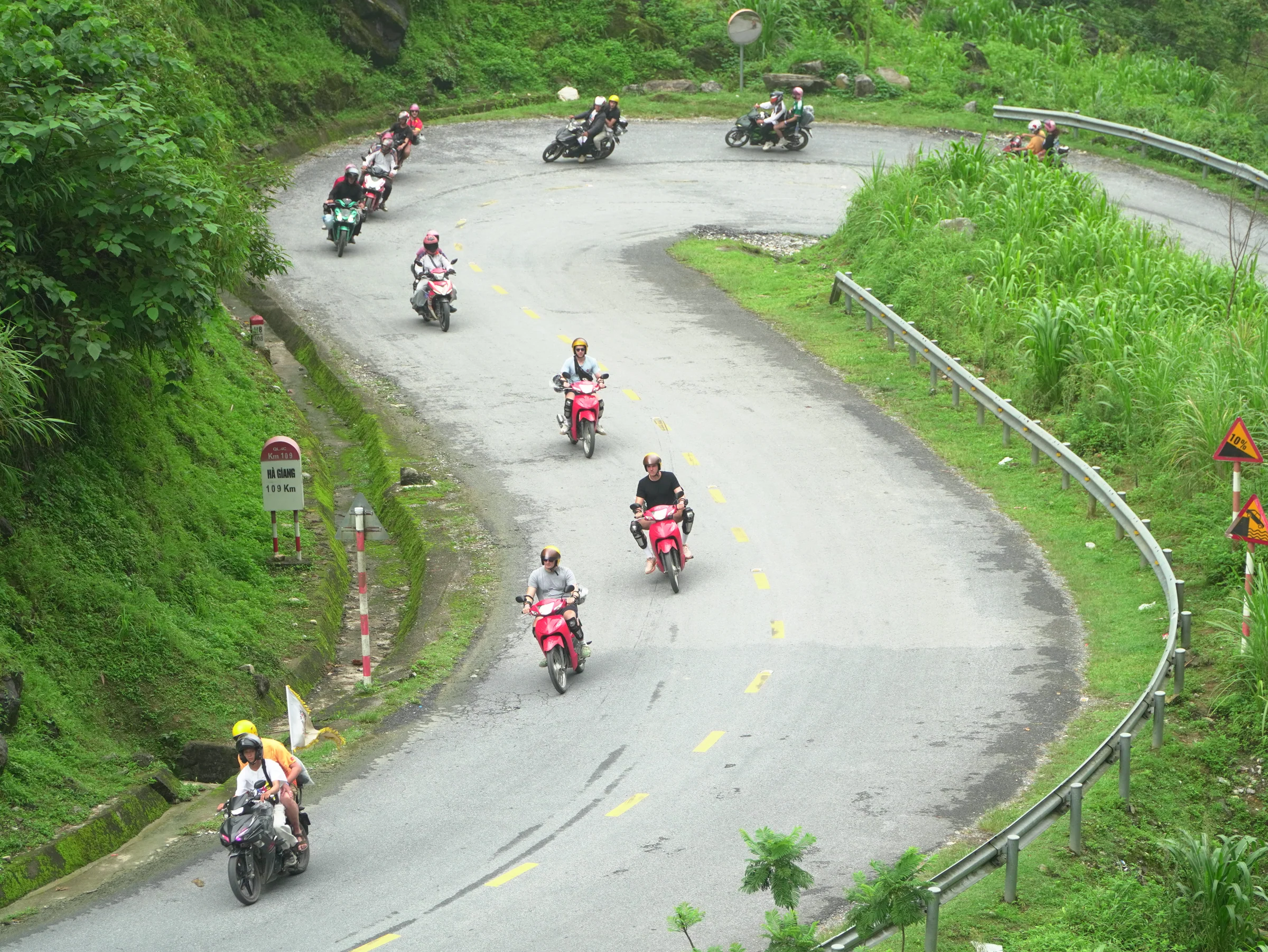 Seven motorbikes strung out along a hairpin bend on a wet mountain road, seen from above. Riders in helmets and knee guards, green karst rising on both sides of the tarmac.