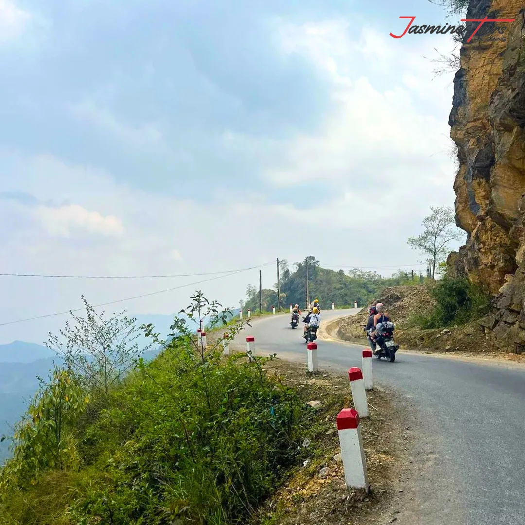 Narrow sealed road on the Ha Giang Loop cut into a limestone cliff, red-and-white edge marker posts along the drop side, three motorbikes riding away in the distance