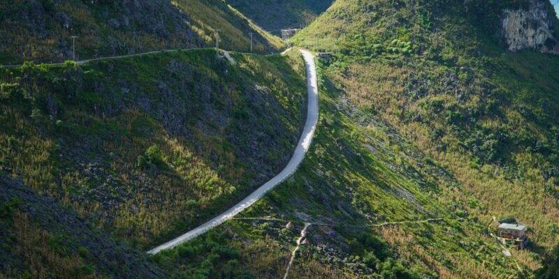 A narrow pale road cut into the flank of a steep grey limestone ridge, running level across the rock face with a near-vertical drop into a deep valley below it and bare stone rising directly above the carriageway.