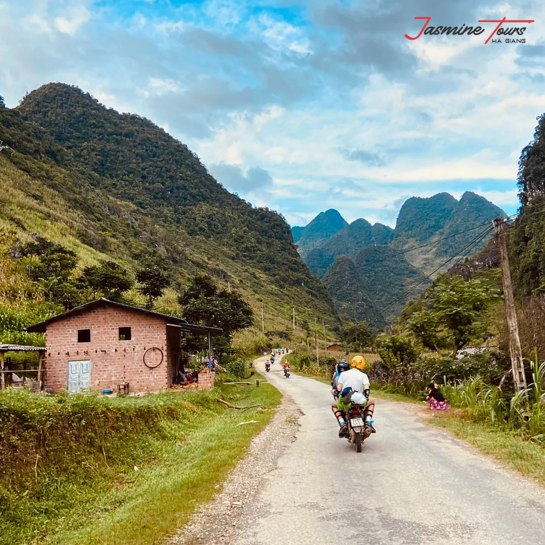 Two riders sharing one motorbike on a quiet single-lane road through a Ha Giang valley, seen from behind, with more bikes ahead