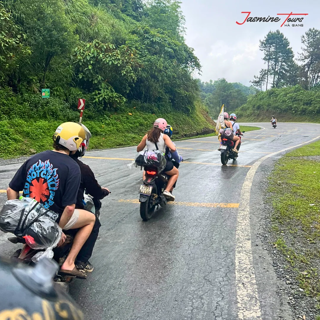 Motorbikes riding in single file on a wet, rain-soaked mountain road on the Ha Giang Loop, spray on the tarmac and low cloud over the trees