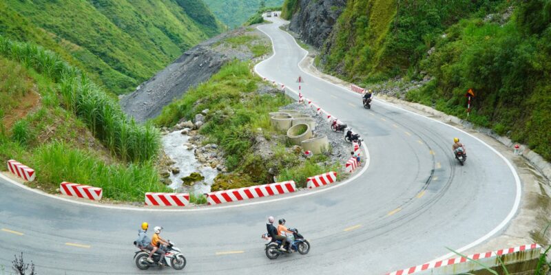 A hairpin bend edged with red and white striped concrete barriers, four small motorbikes working around it, two of them carrying a rider and a helmeted pillion passenger, every rider seen from behind, with a stream running under the road on the inside of the bend.