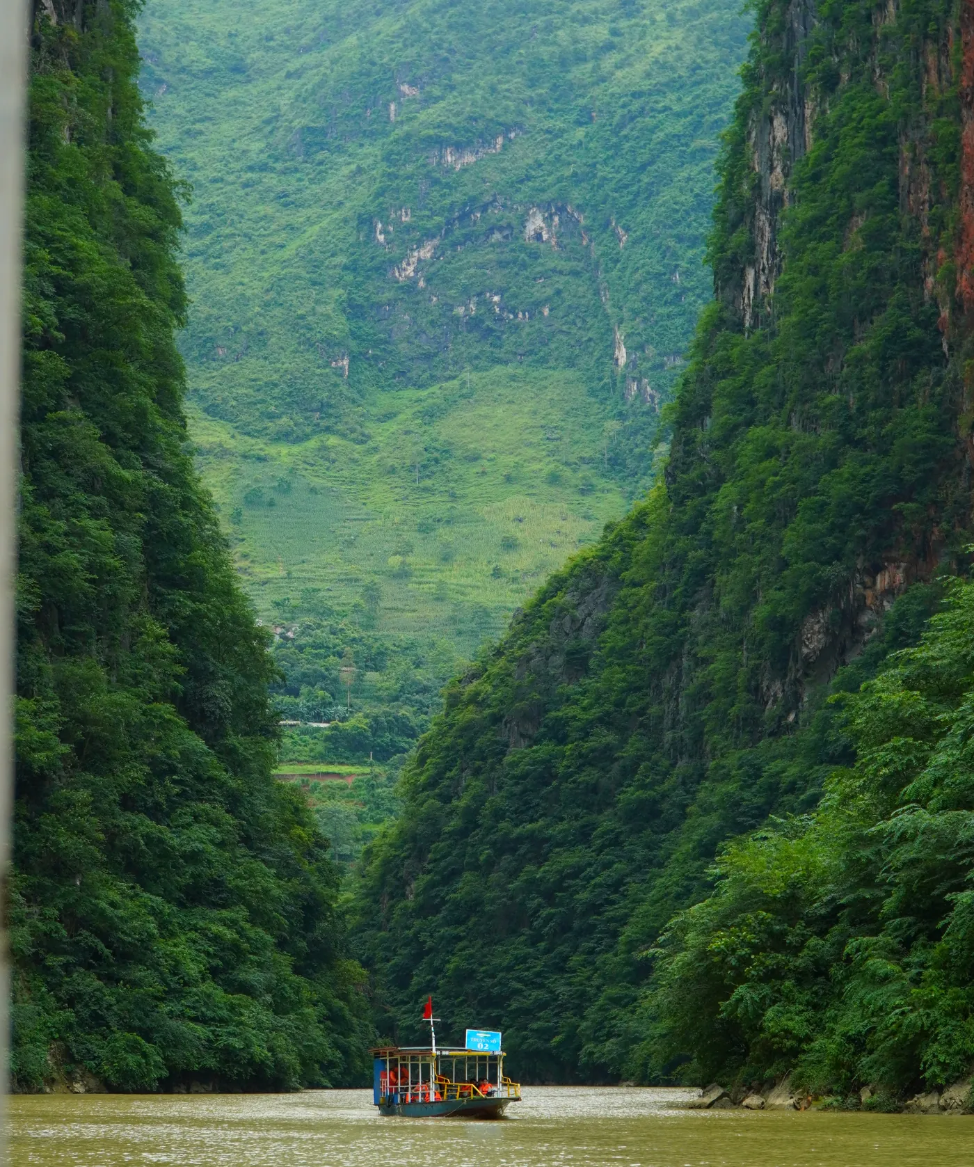 Steep forested gorge walls closing in on a narrow green river far below, with a small covered tour boat on the water and a settlement visible on the valley floor beyond