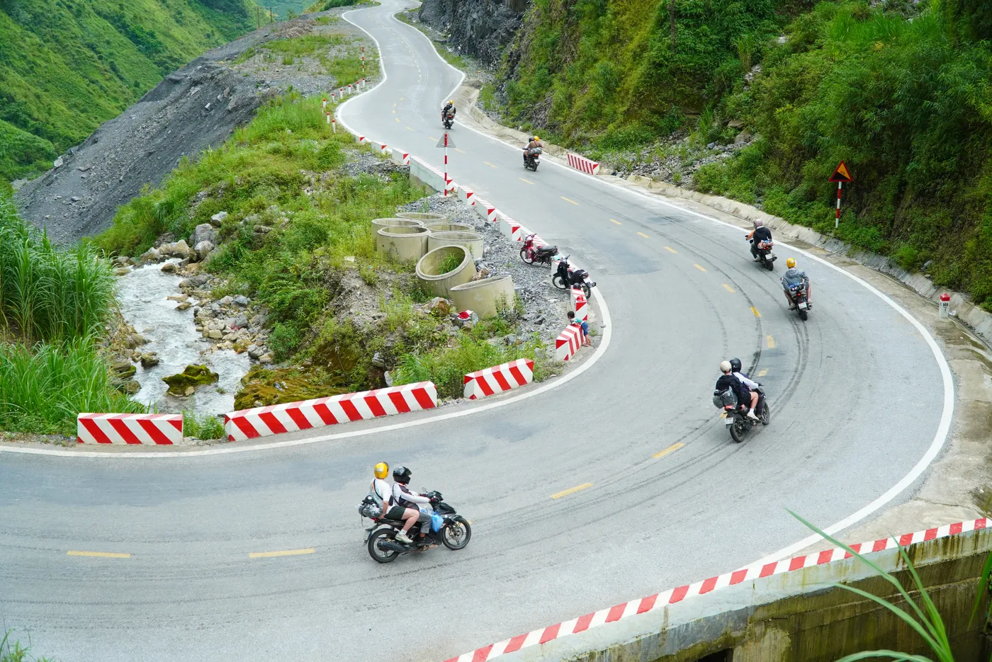 A wide hairpin bend on a mountain road on the Ha Giang Loop with red and white painted kerbing on the outside of the curve and several motorbikes taking the corner