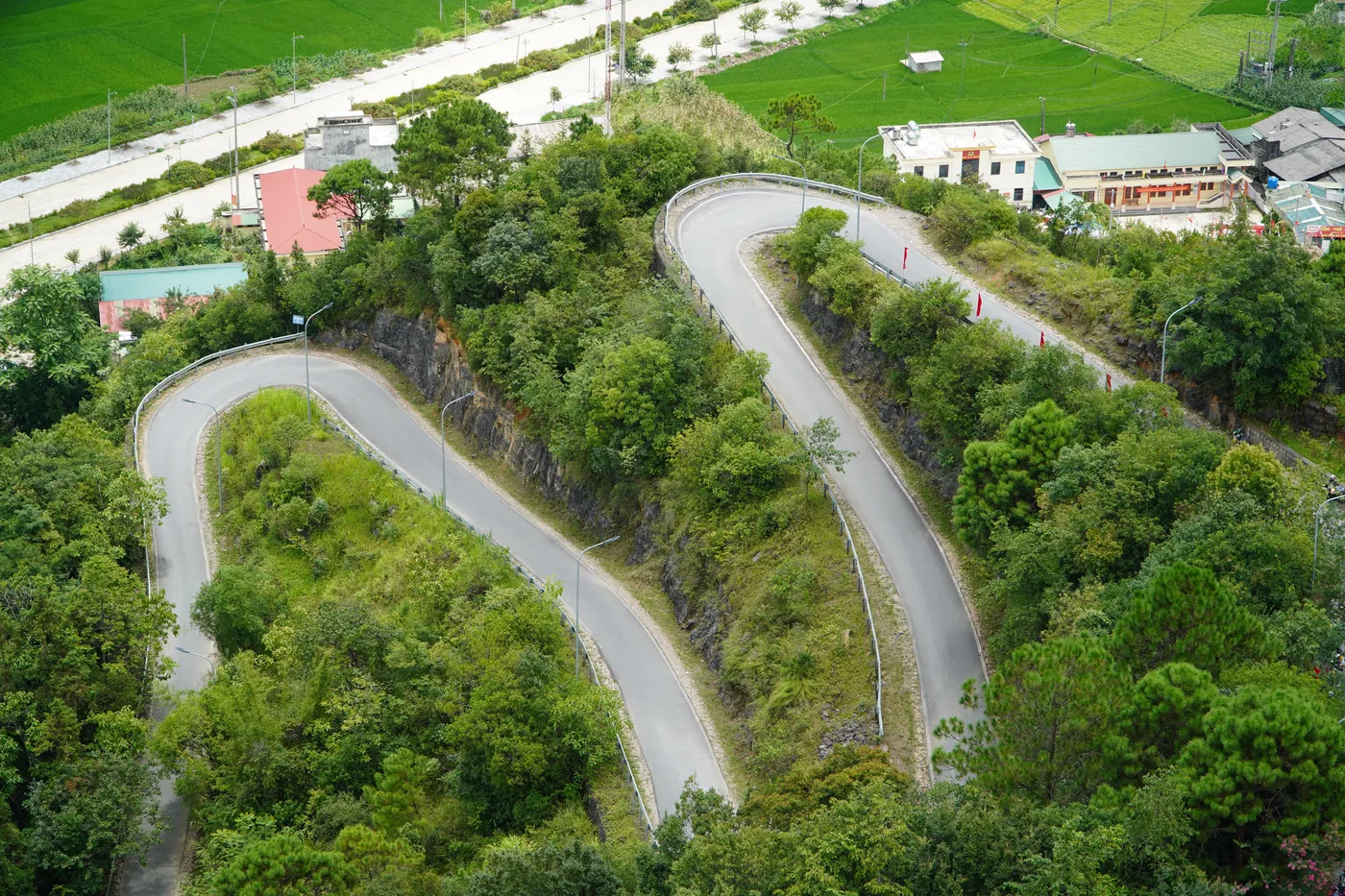 Looking down on a series of stacked hairpin bends climbing out of a valley town on the Ha Giang Loop, with rice fields and houses on the valley floor beyond the bends