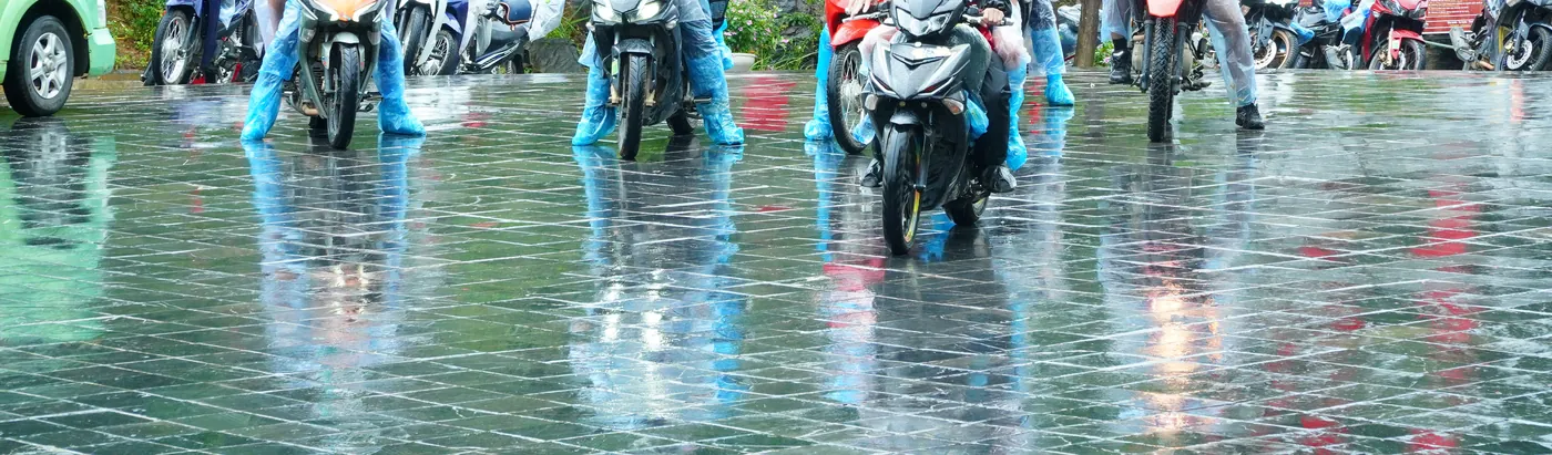 A row of motorbikes on a soaked road surface in Ha Giang, seen low from the front so that only the wheels and the riders' plastic rain-poncho legs are in frame, with the bikes and their colours mirrored in standing water across the tarmac