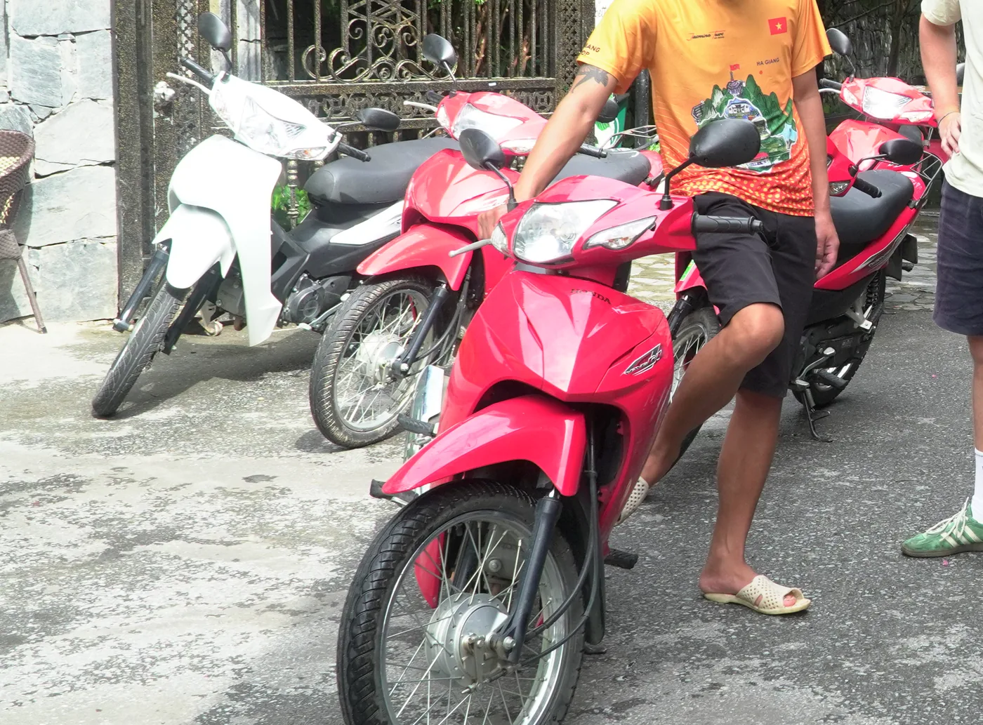 A row of small motorbikes parked on a grey concrete yard beside a stone wall and an ornate wrought-iron gate. The nearest is a bright red Honda semi-automatic with wing mirrors and a black seat; behind it a white scooter with its headlight wrapped in plastic, a black scooter, and a further red bike. A person in an orange T-shirt printed HA GIANG stands beside them, shown from the chin down, in shorts and plastic sandals
