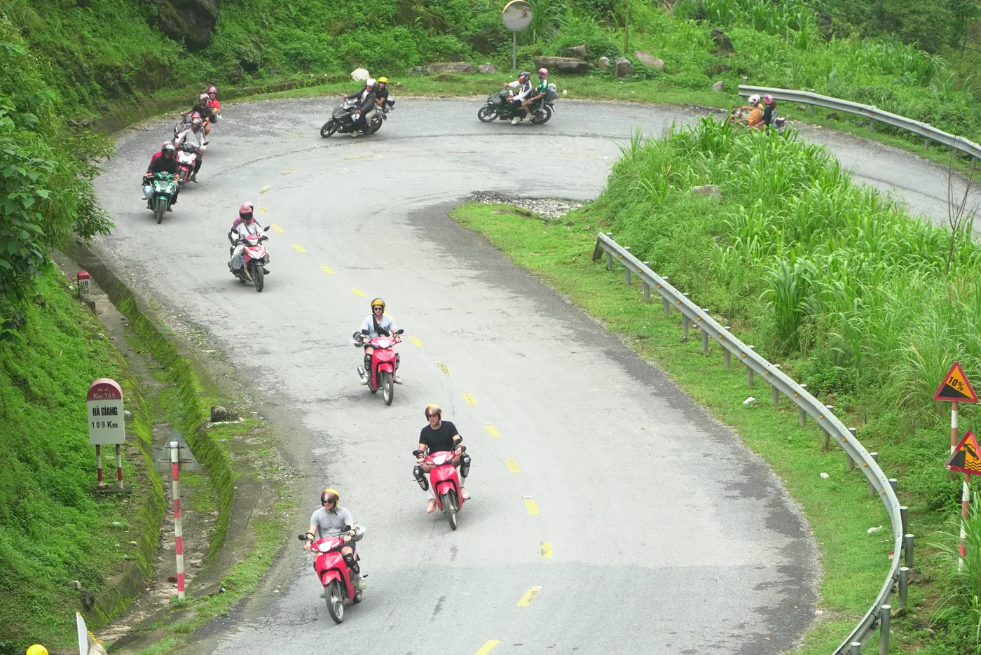 Looking down on a wide hairpin bend on a mountain road, with about ten small motorbikes strung out through the turn in a loose column, every rider helmeted and several in knee pads, thick green vegetation on the uphill side, a steel crash barrier along the outer edge, a red-and-white kilometre marker post at the left reading QL4C, Km 109, HA GIANG 109 Km, and a yellow warning sign for a ten percent gradient at the right