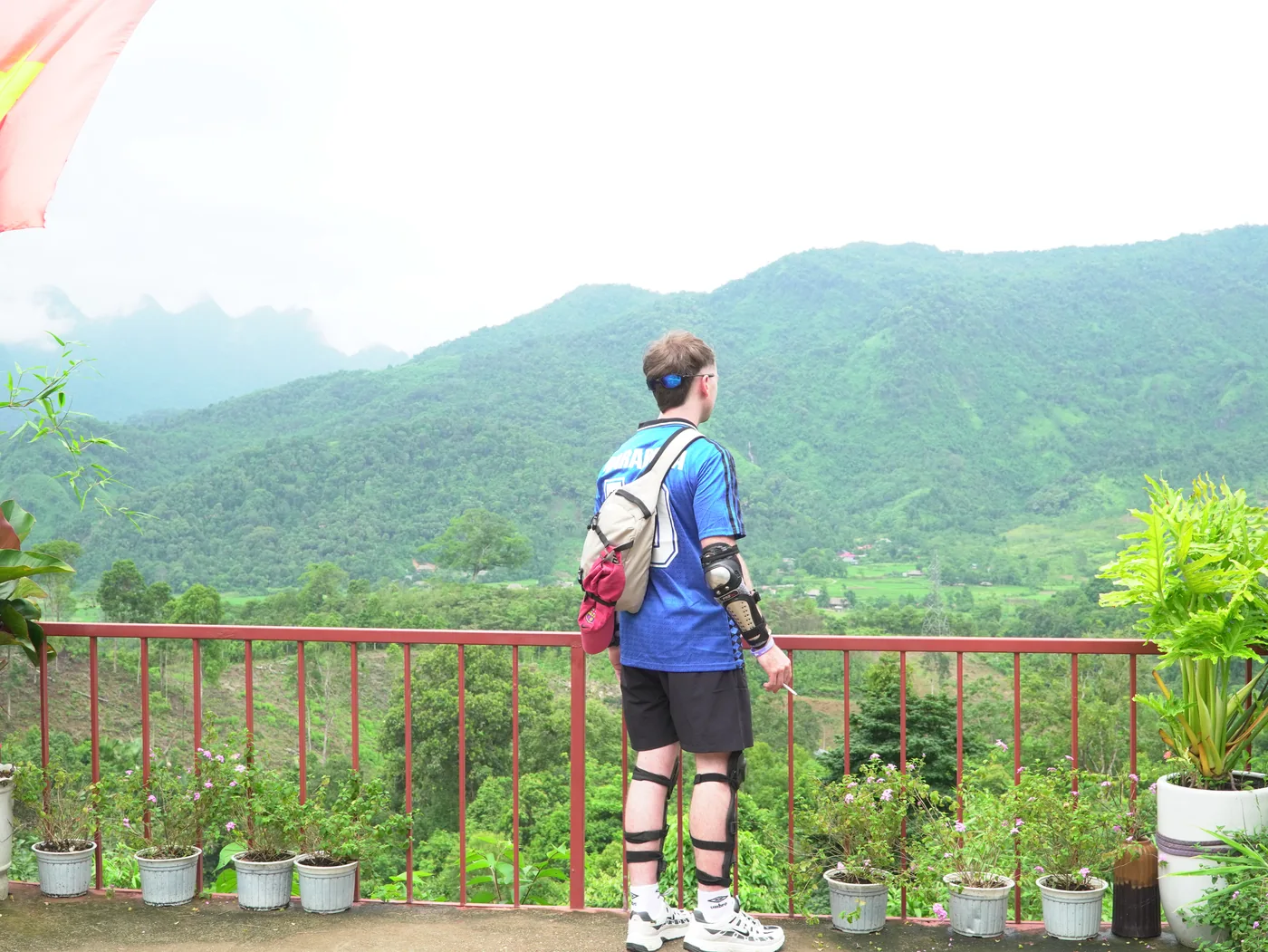 A person seen from behind in a blue shirt and dark shorts, wearing strapped elbow and knee pads and a small waist bag, standing at a red-painted metal railing and looking out over a wide green valley of forested hills under a pale hazy sky. Potted plants line the balcony edge and the corner of a red flag hangs into the top left of the frame