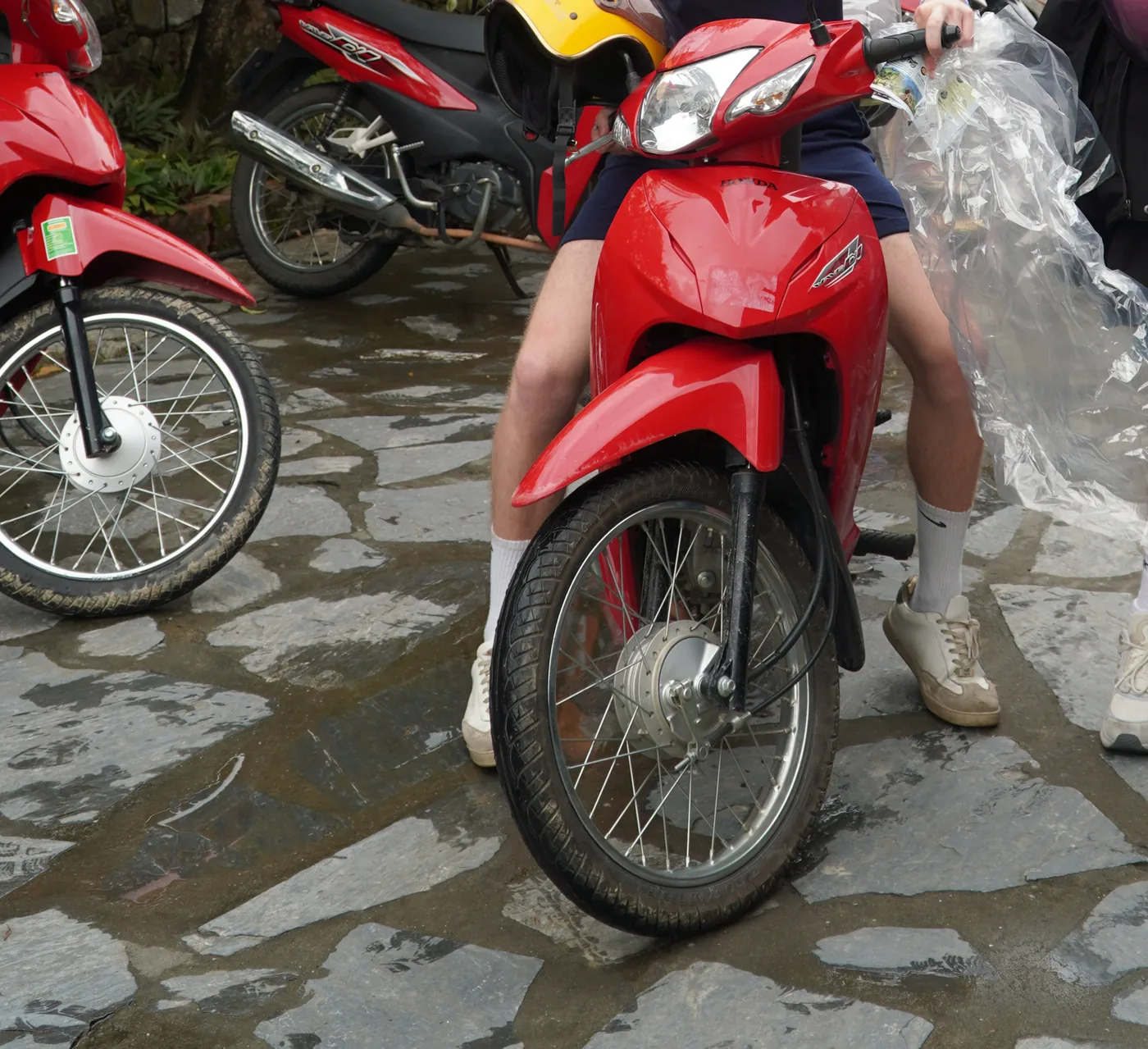 Close view of a bright red Honda semi-automatic motorbike standing on wet, uneven stone paving, its front wheel and mudguard filling the frame. A person's bare legs straddle it and a hand at the right holds a crumpled clear plastic poncho. Behind it another red bike carries a yellow and red open-face helmet hooked over the frame, and a third red bike stands at the left. Water is lying between the paving stones