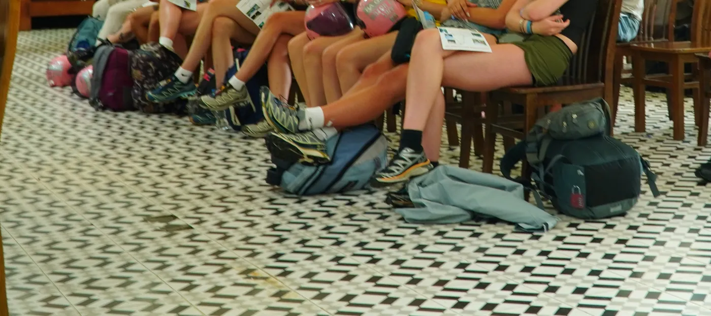 Photographed from the thighs down, a row of seated travellers with backpacks, a soft duffel bag and motorbike helmets on a patterned tiled floor at a morning briefing, printed leaflets resting on their laps