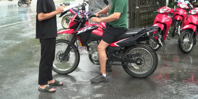 A row of red Honda motorbikes standing on a wet forecourt in the rain, one traveller in shorts and trainers sitting astride a red trail bike while a second person stands beside it holding a clipboard, standing water reflecting the bikes