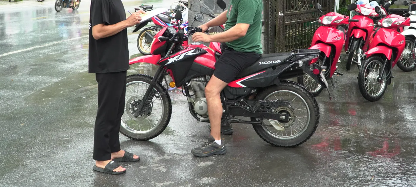 A row of red Honda motorbikes standing on a wet forecourt in the rain, one traveller in shorts and trainers sitting astride a red trail bike while a second person stands beside it holding a clipboard, standing water reflecting the bikes