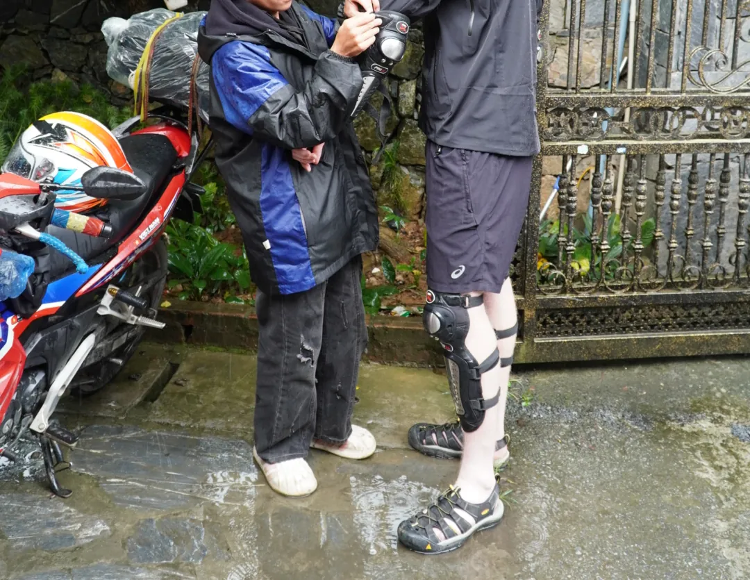 Photographed from the shoulders down, one person in a wet blue and black rain jacket fastens a hinged plastic knee guard onto a second person who is wearing shorts and open walking sandals, on wet stone ground beside a motorbike carrying helmets and a bag wrapped in plastic sheeting