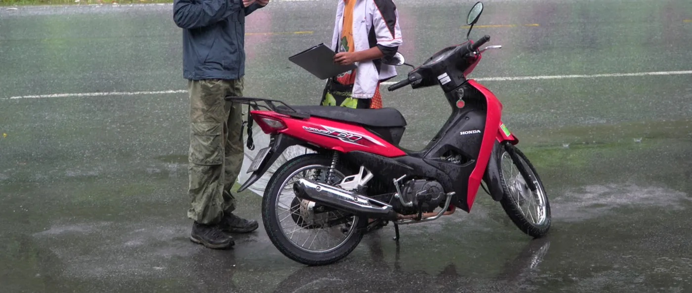 A traveller in a dark rain jacket and cargo trousers stands on a wet road holding a small red document while a second person holds a clipboard, a red Honda Wave step-through motorbike parked beside them in visible rain