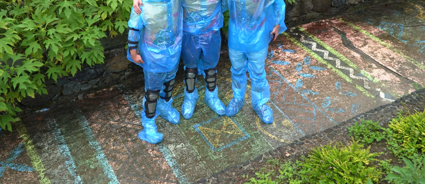 Three people photographed from the chest down wearing thin blue disposable plastic rain suits with matching plastic overshoes, black hinged knee guards strapped over the plastic on two of them, standing on wet painted concrete