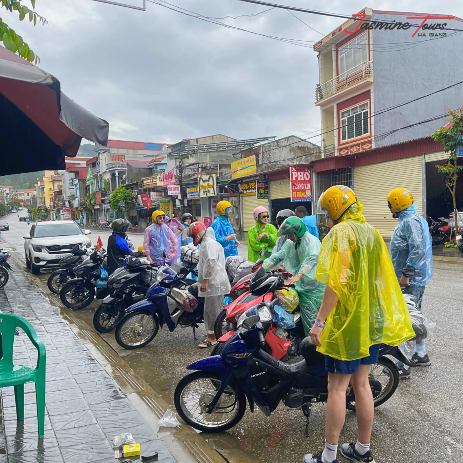 Riders in plastic rain ponchos standing beside parked motorbikes on a wet street in a Ha Giang town during rain