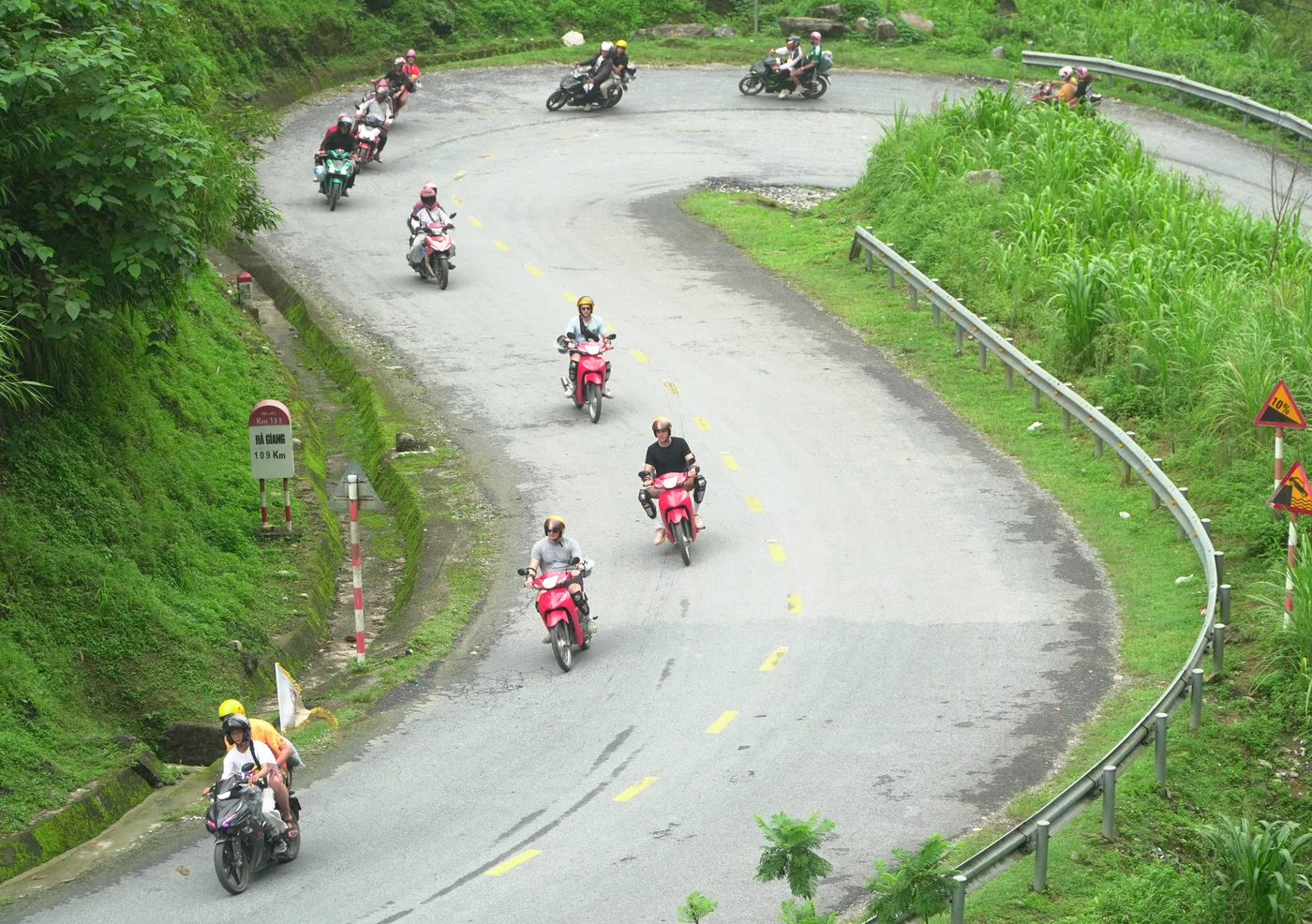 A hairpin bend on highway QL4C in Hà Giang seen from above, with eight small motorbikes strung around the curve. A red-topped kilometre stone stands beside the road on the left reading QL4C, Km 109, HÀ GIANG, 109 Km. A steel guardrail follows the outside of the bend, a convex mirror stands near the apex, and warning signs for a 10 per cent gradient and a sharp curve stand at the right. The asphalt is patched and carries a yellow dashed centre line. Every rider is helmeted and seen at a distance