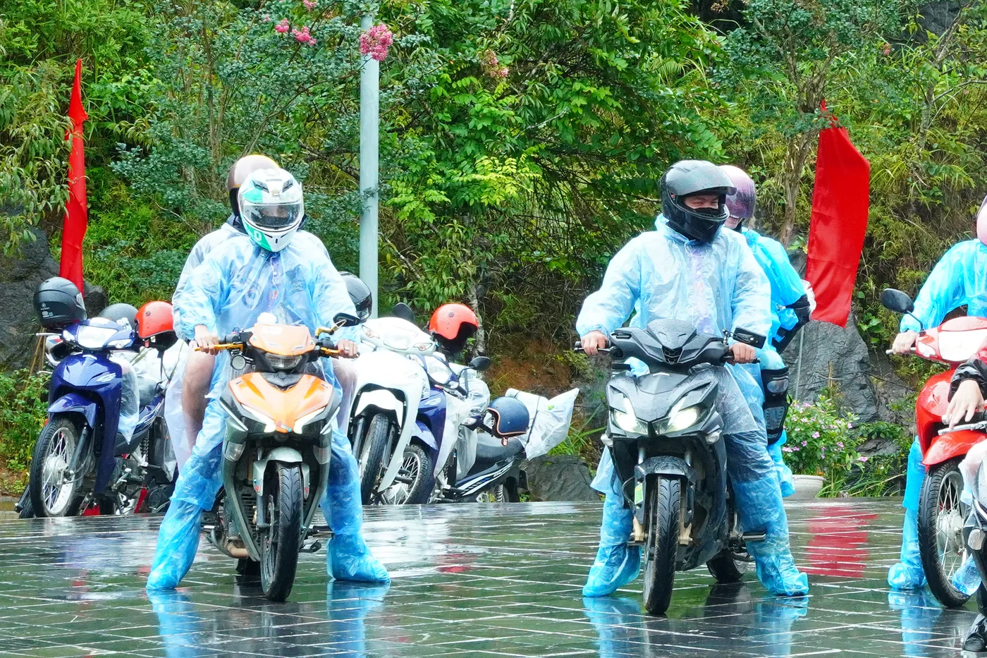 Riders in disposable blue rain ponchos setting off across a soaked paved forecourt, the wet stone reflecting red flags and the bikes above it. The two nearest riders wear full-face helmets with the visors down and no face is legible; blue plastic covers are pulled over their shoes and one machine has its headlight on in daylight. More bikes stand parked along a bank of wet green vegetation behind them