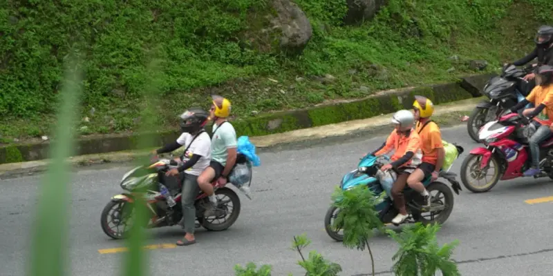 Four motorbikes carrying riders and pillion passengers along a two-lane mountain road in Hà Giang, all helmeted and seen side-on at a distance. A steep green bank rises above the road and a concrete drainage channel runs along its foot. Out-of-focus foliage fills the left edge and the bottom of the frame, a metal guardrail runs along the far side, and the rightmost bike is cut by the frame edge