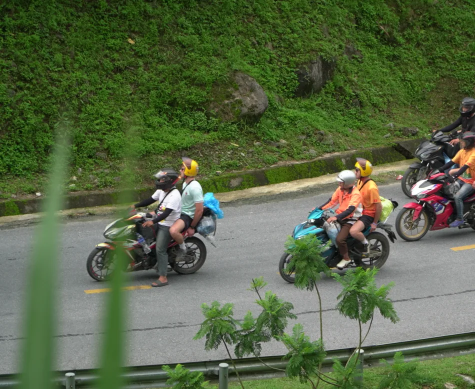 Four motorbikes carrying riders and pillion passengers along a two-lane mountain road in Hà Giang, all helmeted and seen side-on at a distance. A steep green bank rises above the road and a concrete drainage channel runs along its foot. Out-of-focus foliage fills the left edge and the bottom of the frame, a metal guardrail runs along the far side, and the rightmost bike is cut by the frame edge