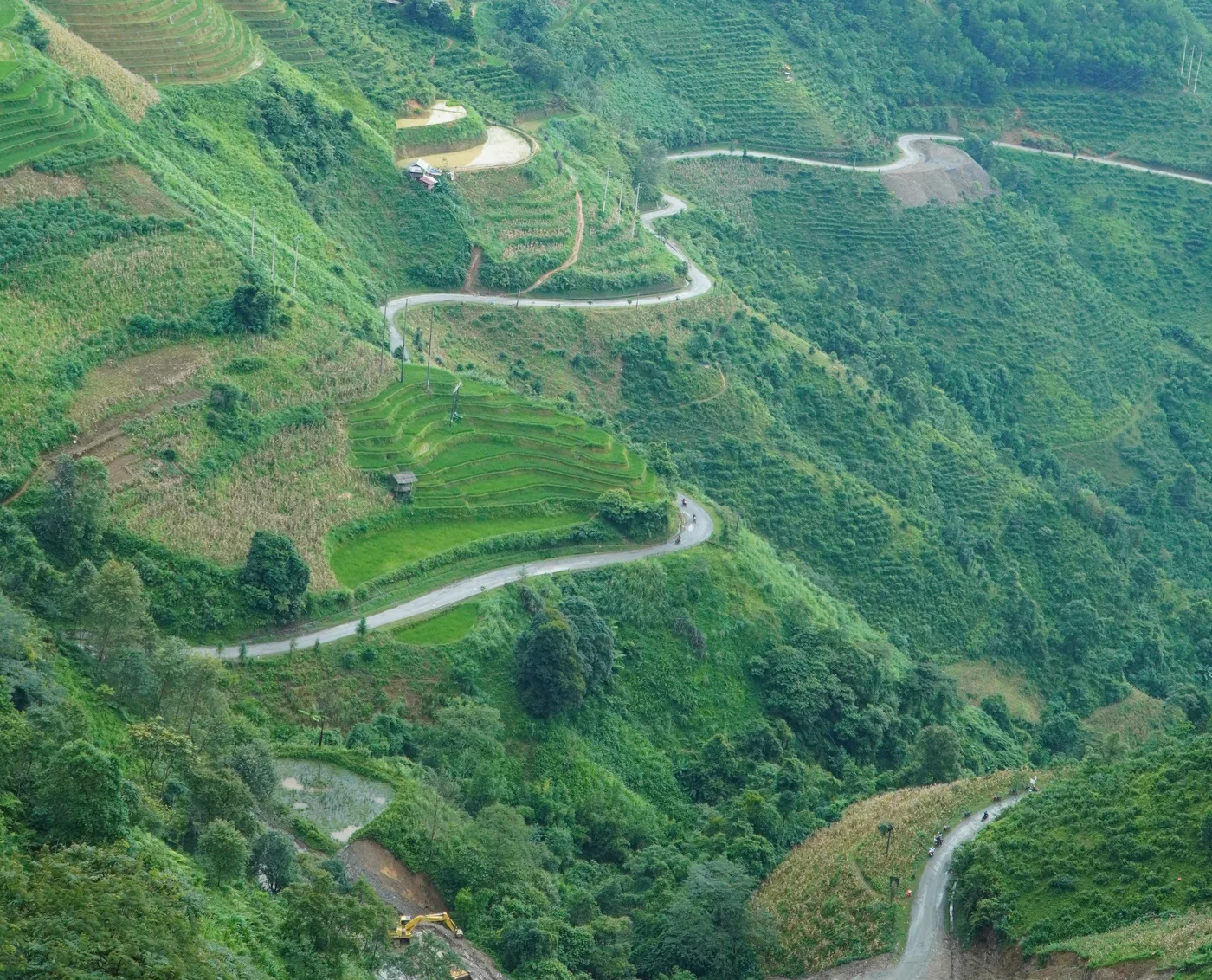 A narrow road switchbacking down a steep valley of terraced fields in Hà Giang, photographed from high above. The road doubles back on itself four times between plots of maize and young rice, and two motorbikes are visible on it as specks. At the bottom of the frame a raw landslide scar of bare orange earth cuts the slope, with a yellow excavator standing on cleared ground beside it. No person in the frame is identifiable
