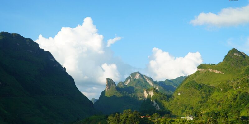 Steep conical limestone peaks rising in ranks under a deep blue sky with white cumulus cloud, the nearer slopes green with vegetation and the valley floor lying in shadow between them.