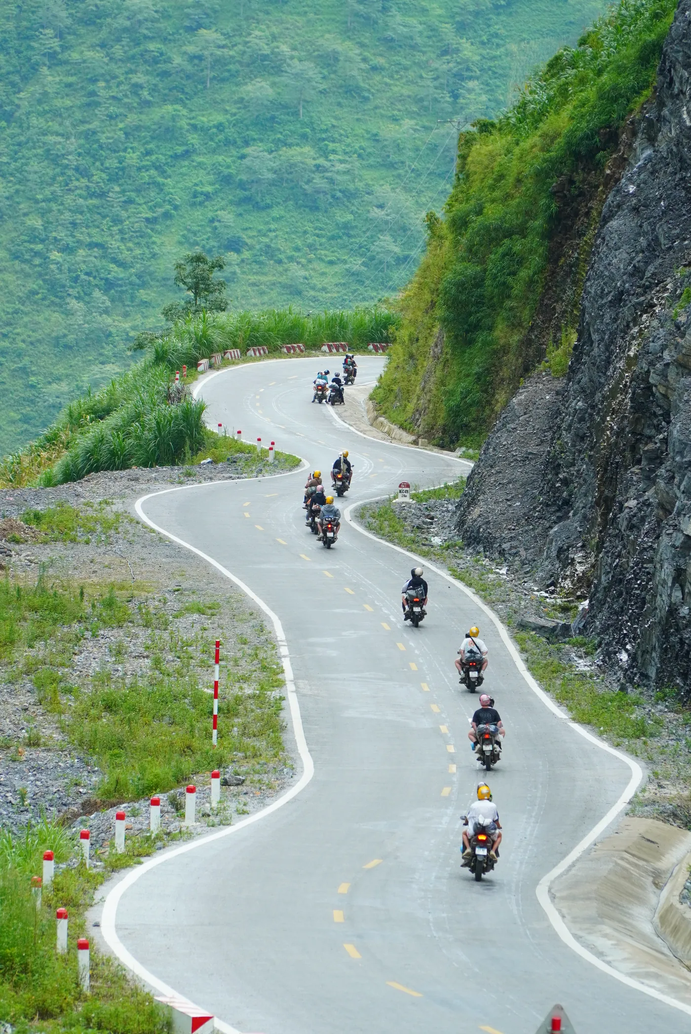A road curving in a tight S-bend along a steep green mountainside in Hà Giang, red and white marker posts on the drop side, with a line of motorbikes riding down through the bends