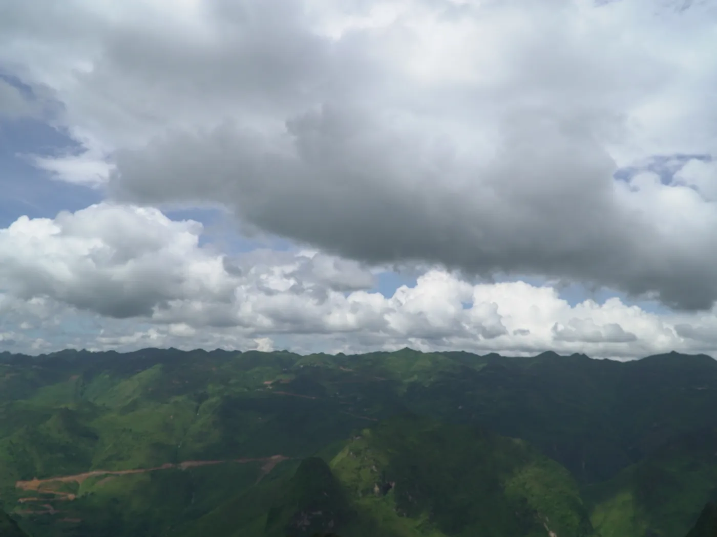A wide view over rolling green ridges in Hà Giang receding to a dark horizon, under a tall sky of heavy broken cloud