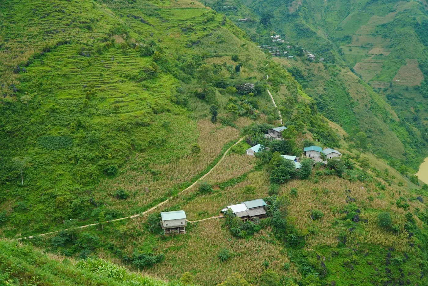 A settlement of small buildings on a broad green hillside in Hà Giang, cultivated terraces stepping down the slope below it and forested ridges rising behind