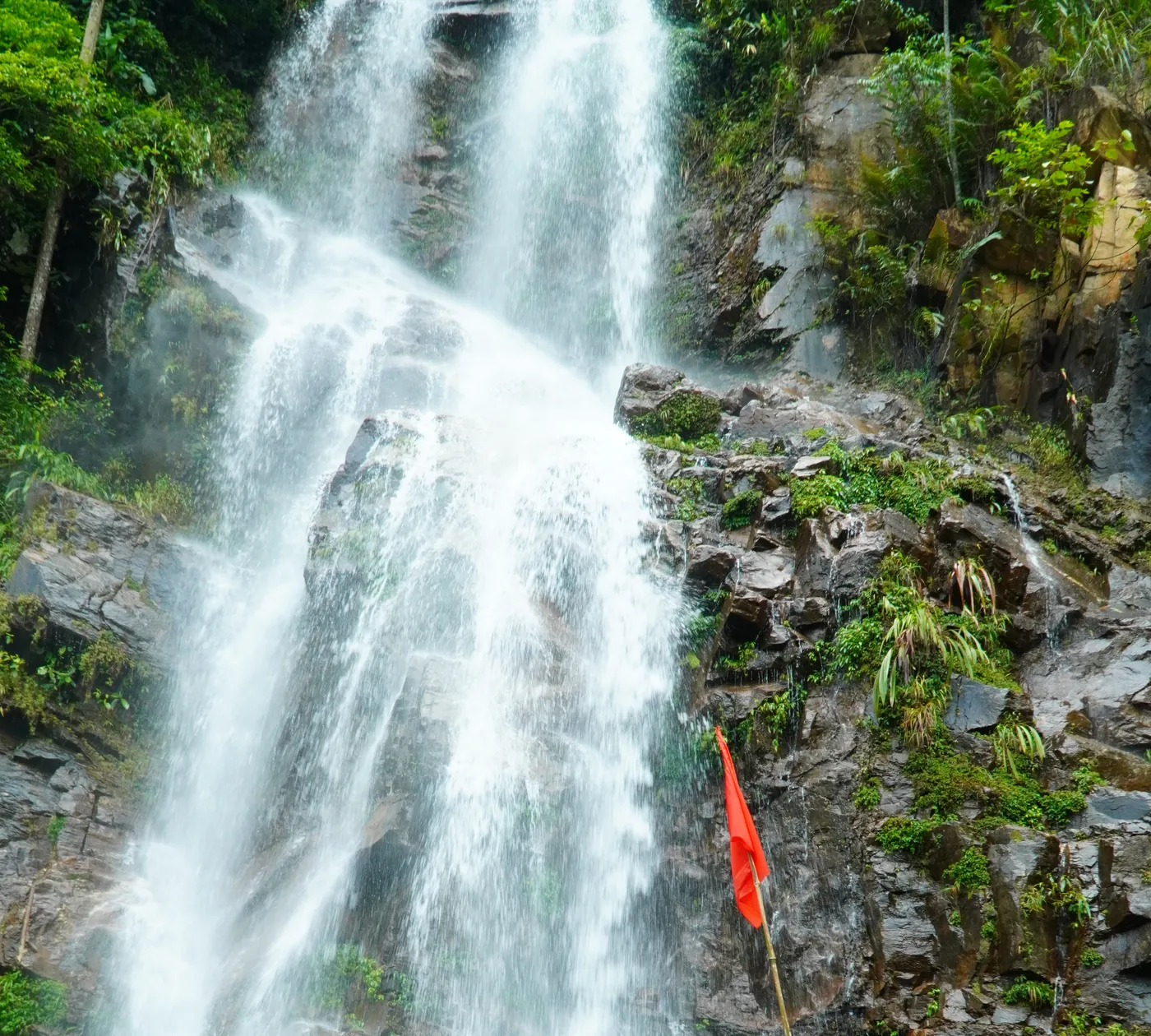 A wide waterfall breaking over dark stepped rock in Hà Giang, several separate falls of white water among wet ledges and dense green vegetation, with a small red flag on a pole at the right