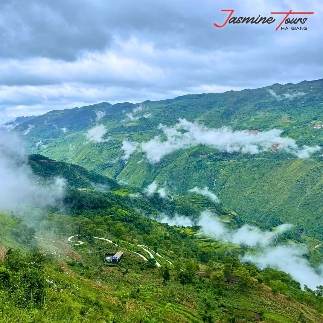 Cloud streaming through a green valley on the Ha Giang Loop, with terraced fields and a pale switchback road visible below the cloud line