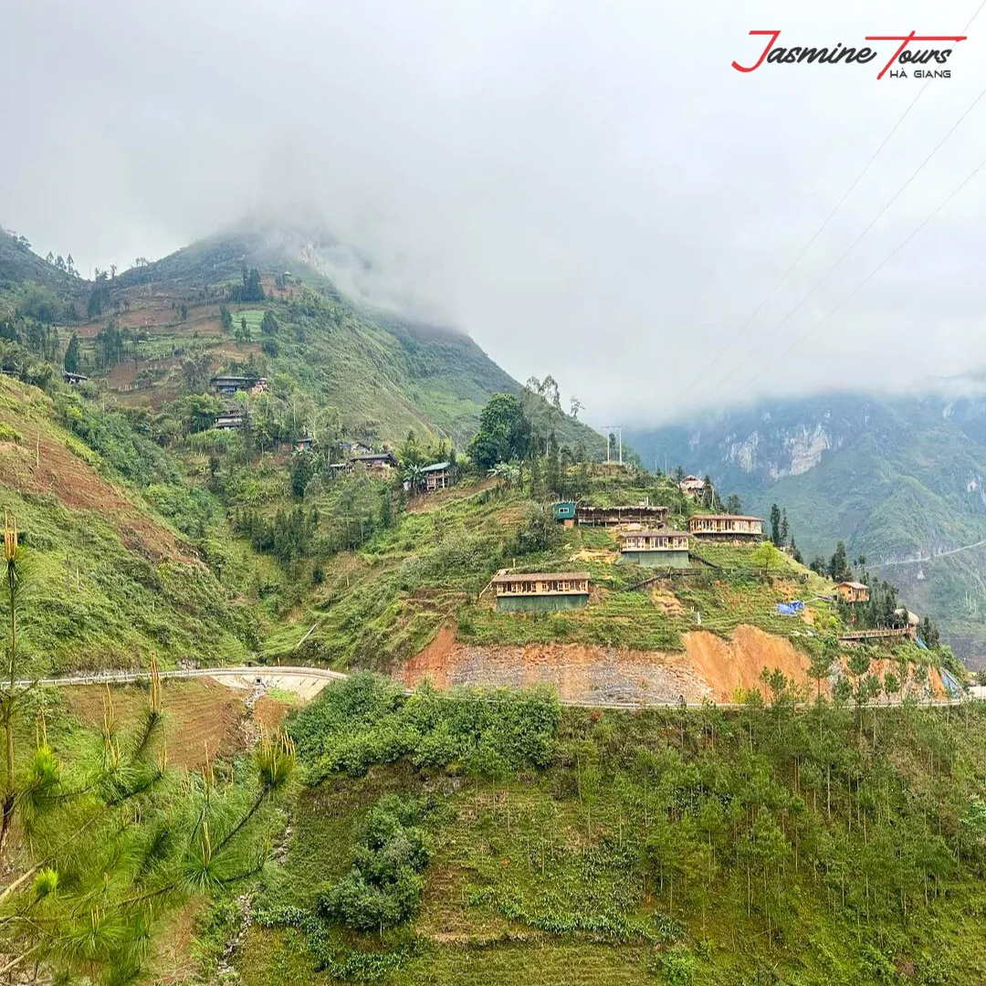 A hillside village on the Ha Giang Loop under low cloud, with bare orange cut earth on the slope above the road