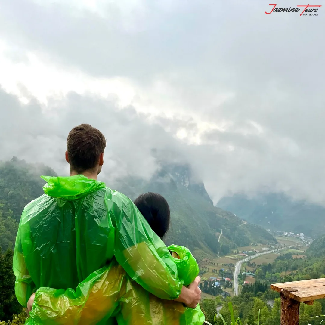 Two riders in green plastic rain ponchos seen from behind at a Ha Giang viewpoint, looking out over a valley filled with low cloud