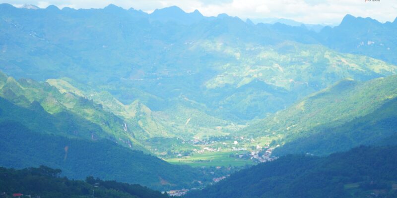 A wide green valley seen from high ground, a scattered small town spread across the valley floor, terraced fields on the near slopes and ranks of forested ridges receding into haze behind it.
