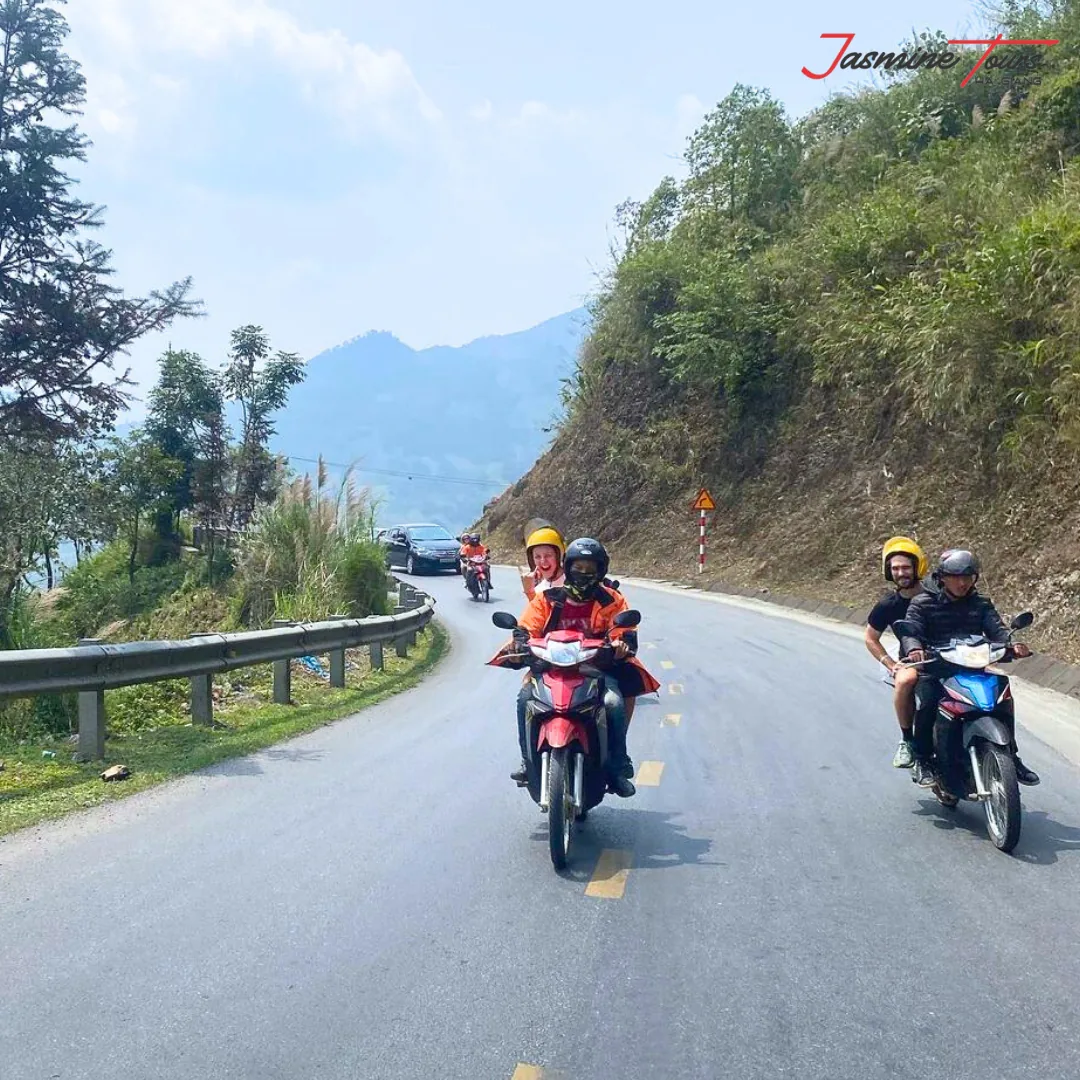 Motorbikes and a car sharing a two-lane sealed highway in northern Vietnam, with a crash barrier on the left and a wooded hillside on the right