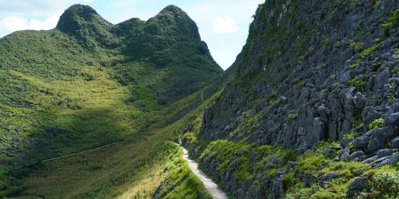 A dark limestone peak rising steeply above a narrow pale road that threads along the base of the rock face, green slopes falling away on both sides and further peaks standing behind it.
