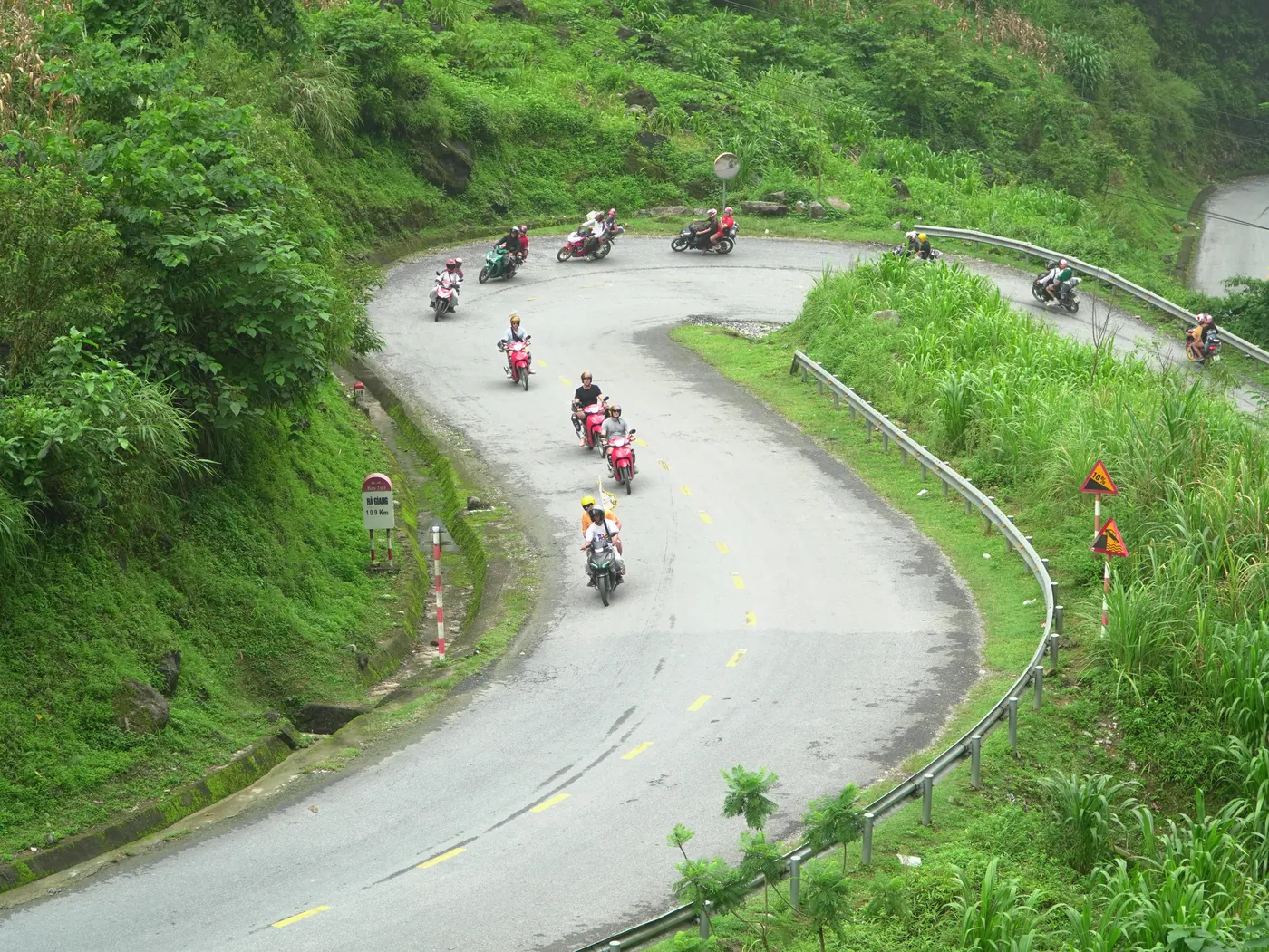 A hairpin bend on a two-lane mountain road seen from above, with about a dozen small motorbikes in helmets strung out through the turn, a steel crash barrier along the outer edge, red-and-white warning signs, and a kilometre marker post at the left reading QL4C, Km 109, Ha Giang 109 Km