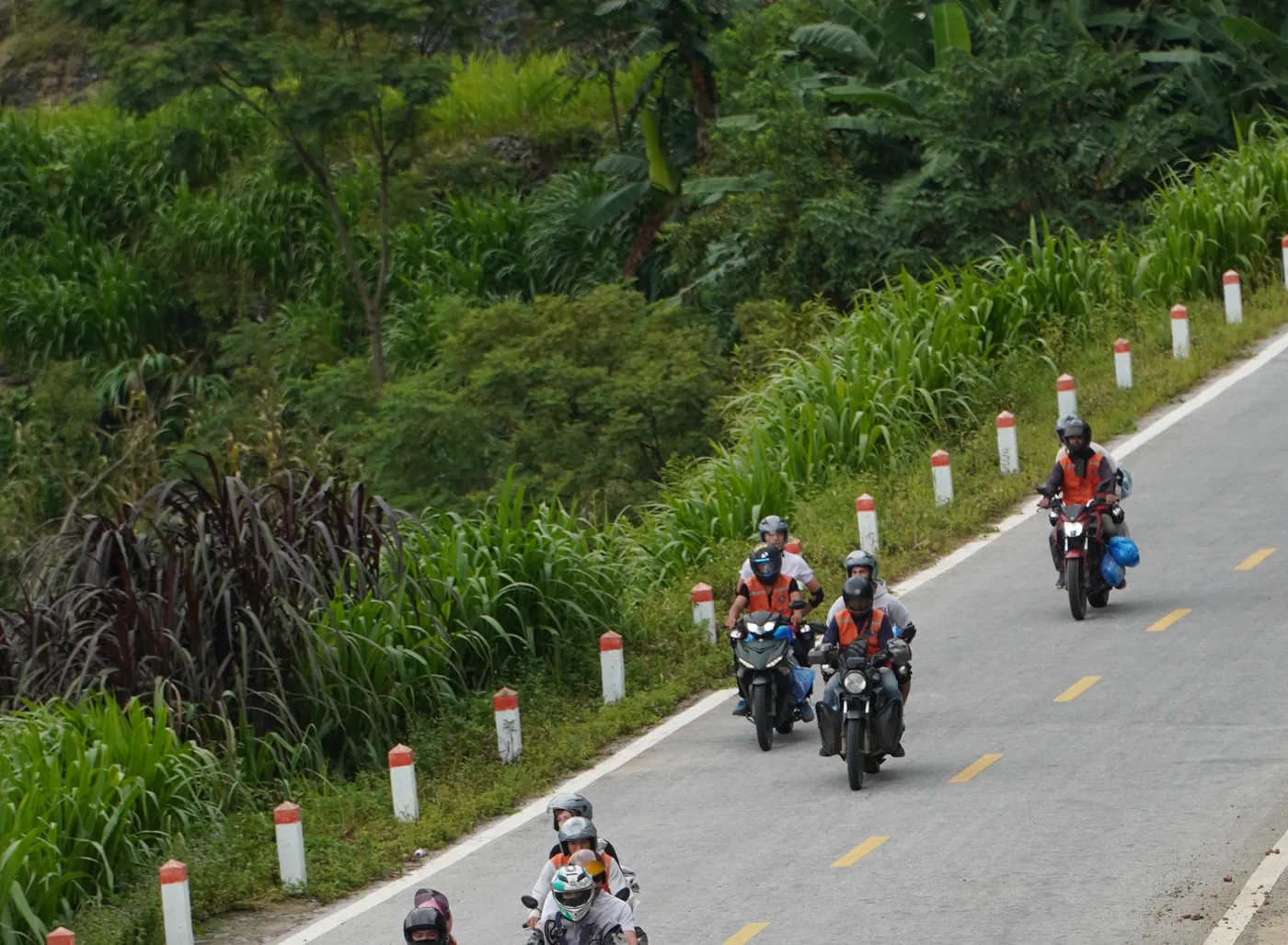 A wide two-lane mountain road with fresh yellow centre markings and a line of red-and-white kerb posts, seen from above, with about eight motorbikes in helmets working downhill in a loose column and dense green vegetation on the uphill side