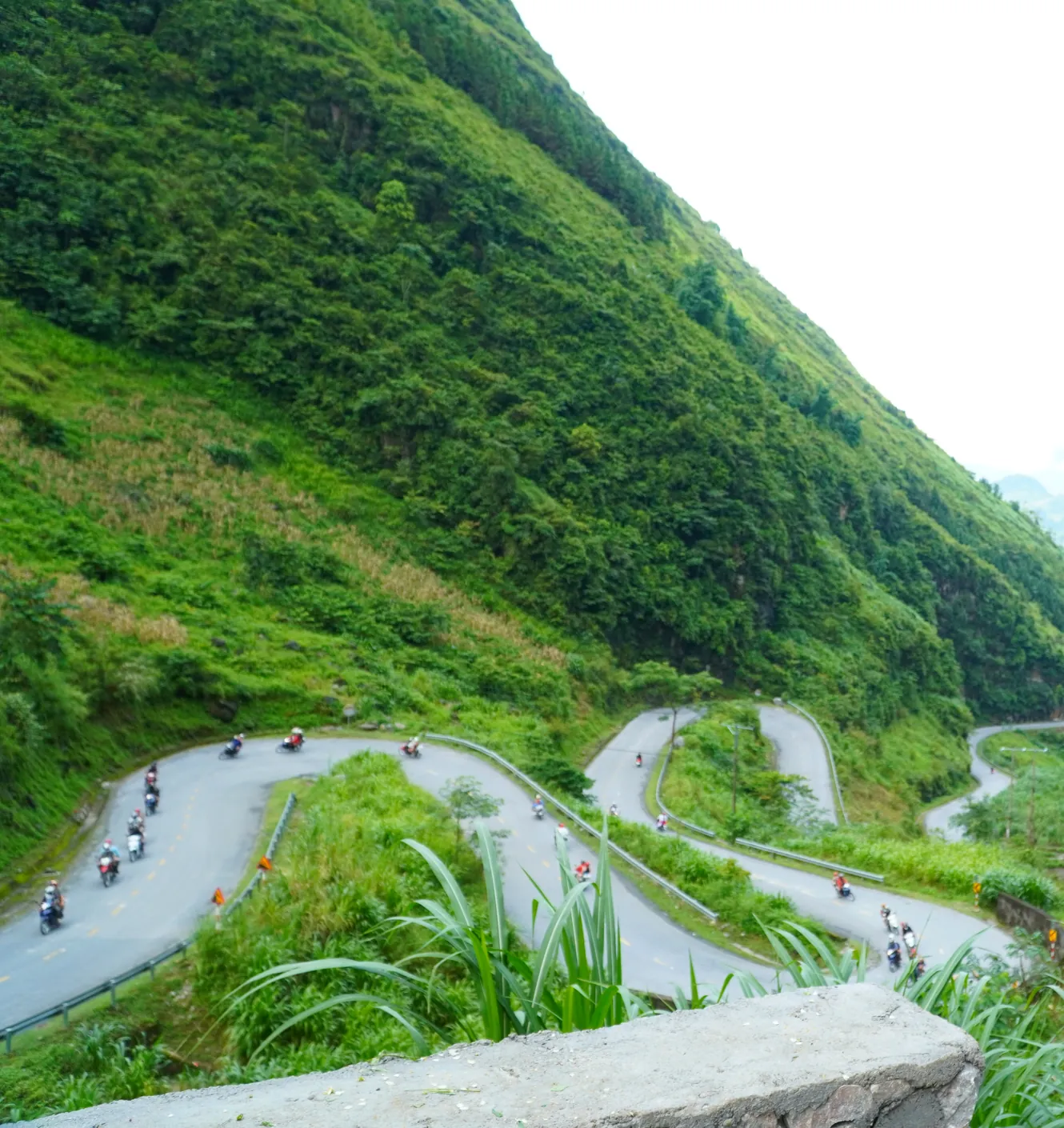 A steep green mountainside seen from above, with a road folding back on itself through four hairpin bends and a scattered line of small motorbikes working down through them, a concrete parapet in the foreground