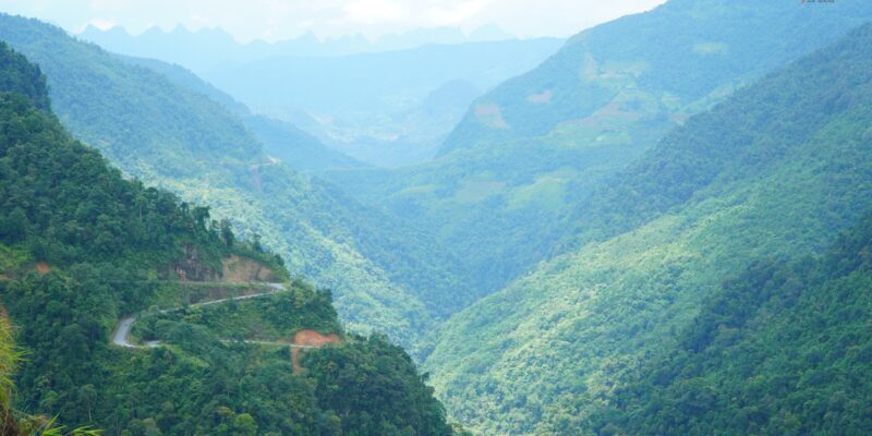 A deep forested gorge seen from high on one side, a road terrace cut into the near slope and doubling back on itself, ranked ridges receding into haze at the head of the valley, and a thin pale line of water on the valley floor far below.